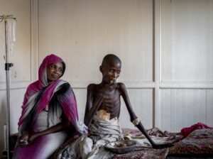 Omdurman, Sudan. Hassan Adam, 14, ate food in his room at Al Nau hospital as his mother Amona Elhadi looked on. Hassan had only recently started to be able to eat again, having been shot by what his mother claimed was a member of the armed civilian resistance known as mustanfareen. Sudan’s military has been mobilising civilians across the country to help fight against RSF forces.